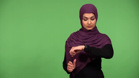 A Young Beautiful Muslim Woman Taps Her Watch and Looks at the Camera  Green Screen Background