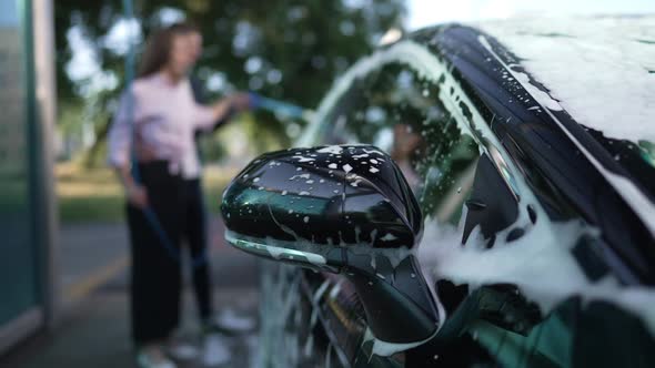 Closeup Black Car Side View Mirror with White Foam and Blurred Couple Washing Vehicle at Background alt