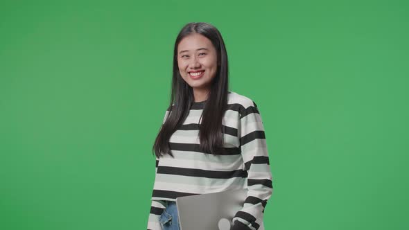 A Smiling Asian Woman Holding Computer While Standing In Front Of Green Screen Background alt