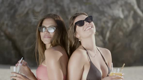 Happy Girls Standing Back to Back Drinking Cocktails on Beach alt