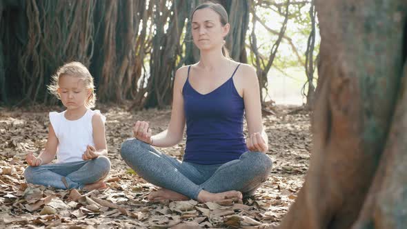 Little Child Girl with Young Mother Meditating Together Under Banyan Tree