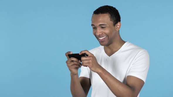 Young African Man Playing Game on Smartphone Blue Background alt