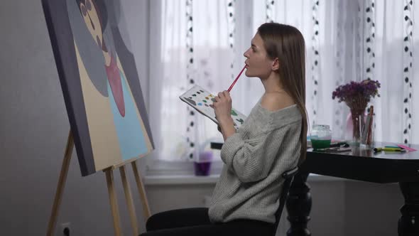 Side View Gorgeous Female Painter Making Strokes with Acrylic Paint Brush Sitting at Picture alt