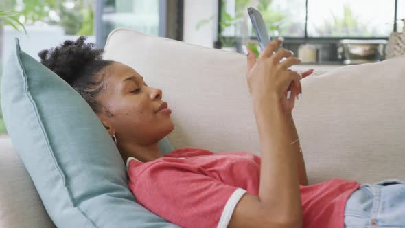 Video of happy african american woman lying on sofa and using smartphone alt