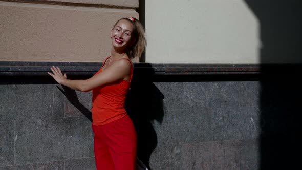 Portrait of a Smiling Blonde in Red Clothes Near the Wall of the Building alt