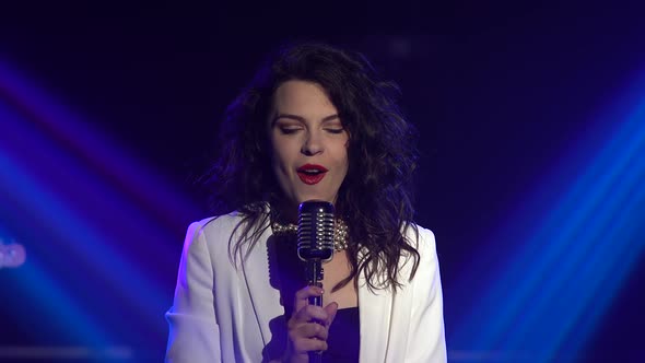 A Stylish Girl Vocalist Sings on Stage in a Vintage Microphone. Dark Studio with Smoke and Neon alt