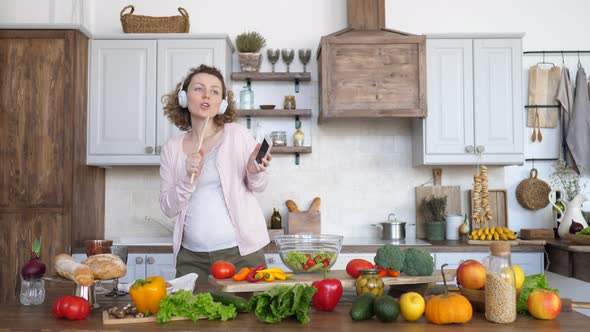 Pregnant Woman Dancing And Singing In Headphones With Mobile Phone On Kitchen. alt