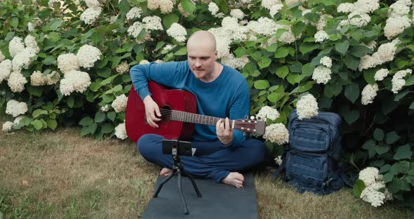 Man with Guitar Sitting Among Flowers and Sings While Reading Text From Phone