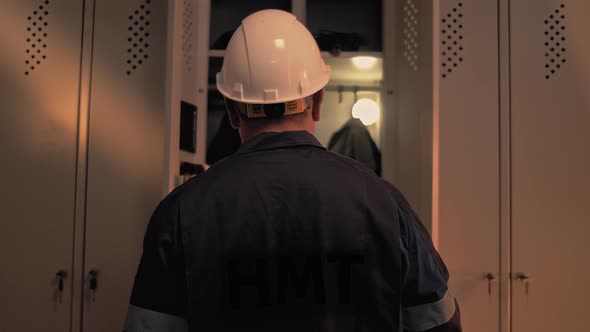 Oil refinery worker stands at his open locker in the locker room and puts a safety helmet