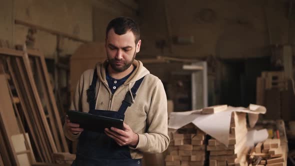 A Young Man with a Beard Walks Around the Carpentry Shop with a Tablet in His Hands alt