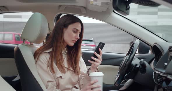 Elegant Woman Sitting in Car and Surfing Feed News on a Smartphone and Drinking Coffee alt