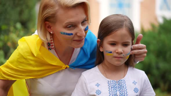 Loving Ukrainian Mother Drawing on Face of Pretty Daughter with National Flag Colors and Looking at alt