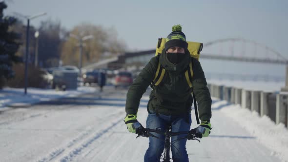 Portrait of Cyclist with Backpack and Protective Mask Rides Bicycle on Snow Covered Street with Food alt