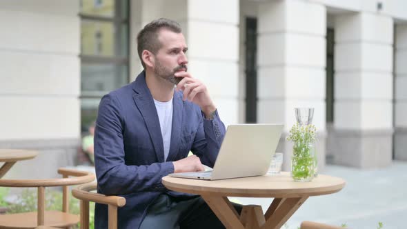 Man Thinking While Using Laptop Sitting in Outdoor Cafe alt