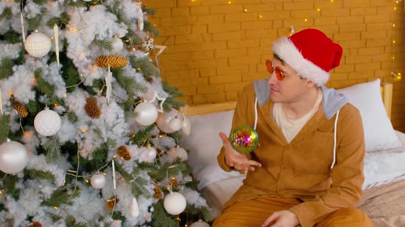 Portrait of Man in Santa Hat and Bright Glasses with Disco Bauble Sitting on Bed Near Christmas Tree alt