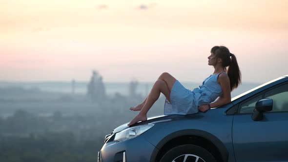 Happy Woman Driver in Blue Summer Dress Enjoying Warm Evening Laying on Her Car Hood alt