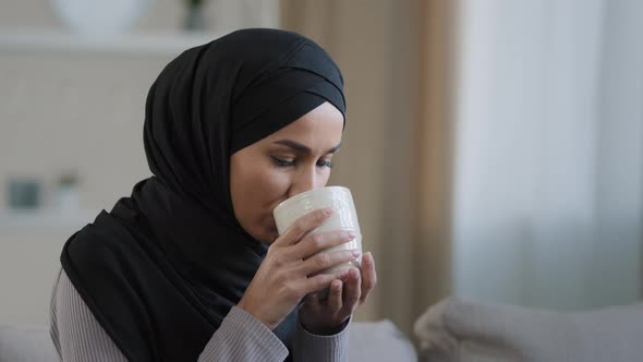 Carefree Joyful Muslim Girl in Hijab Drinking Tea Relaxing in Room Alone Look Thoughtfully Into alt
