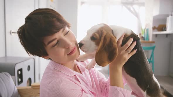 Young Caucasian Female in Pink Pyjamas Holding Beagle Puppy in Arms and Kissing Him in Slowmotion alt
