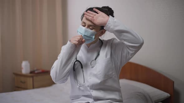 Portrait of Tired Ill Caucasian Young Doctor in Covid19 Face Mask Coughing Touching Forehead Sighing alt