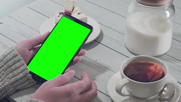 Woman holding smartphone with green screen with hot tea and cake on table