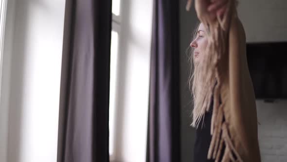 Cheerful Woman Standing Wrapped in Plaid Smiles and Looks at Window at Home alt