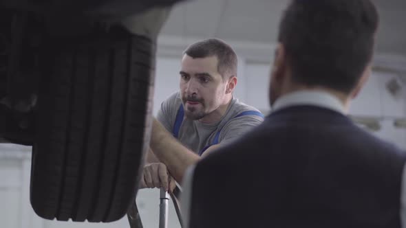 Shooting Over Shoulder of Serious Worker Looking at Car Wheels. Auto Mechanic Checking Fixed alt