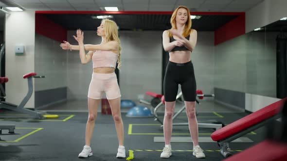 Wide Shot Portrait of Two Confident Young Caucasian Women Stretching Hands Training in Gym alt
