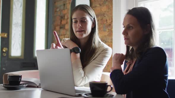 Two smiling caucasian businesswomen sitting at table with coffee, using laptop and talking, alt