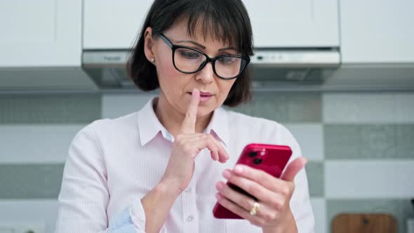 Mature Serious Woman Looking Reading in Smartphone Screen Kitchen at Home alt