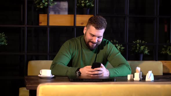 Happy Bearded Man Sitting in a Cafe and Using a Smartphone
