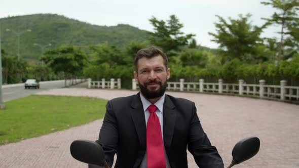 Portrait of a Senior Bearded Smiling Businessman Driving Up on a Motorcycle Along the Street alt