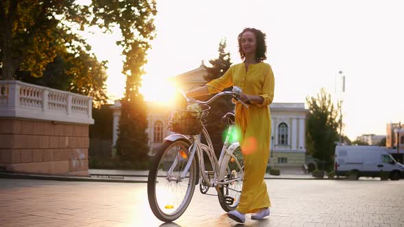 Beautiful Young Woman in a Long Yellow Dress Walking During the Dawn Holding Her City Bicycle's alt