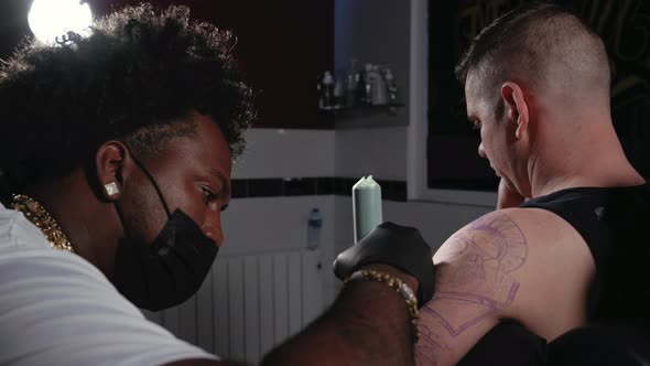 Tattoo Artist is Performing His Work on a Young Man in His Studio alt