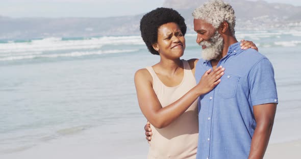 Happy african american couple walking and embracing on sunny beach alt
