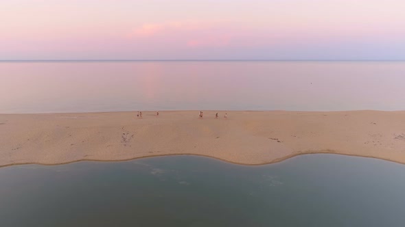 Group of People Walking on Sand Stripe at Sea Coast Against Flat Sea Horizon at Sunset with Pink alt