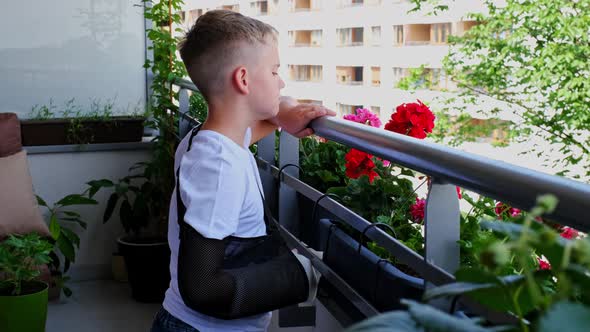 Sad Child with a Broken Arm in a Cast Looks Out Into Yard From the Balcony alt