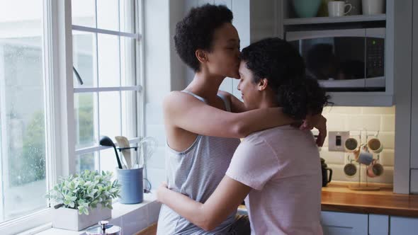 Mixed race lesbian couple hugging and drinking coffee in kitchen alt
