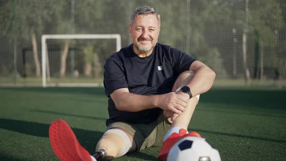Portrait of Confident Smiling Handsome Sportsman Amputee Sitting on Summer Stadium Looking at Camera alt