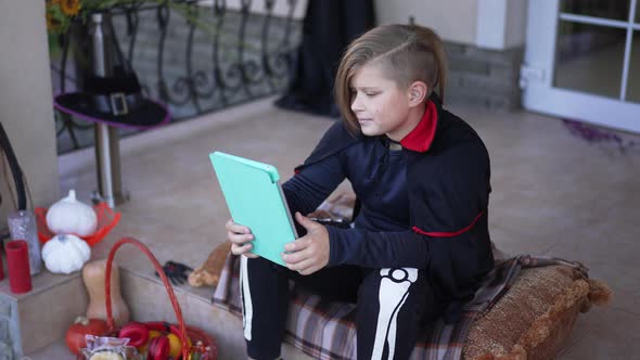 Cute Boy in Vampire Skeleton Costume Watching Movie Online on Tablet Smiling in Slow Motion Sitting alt