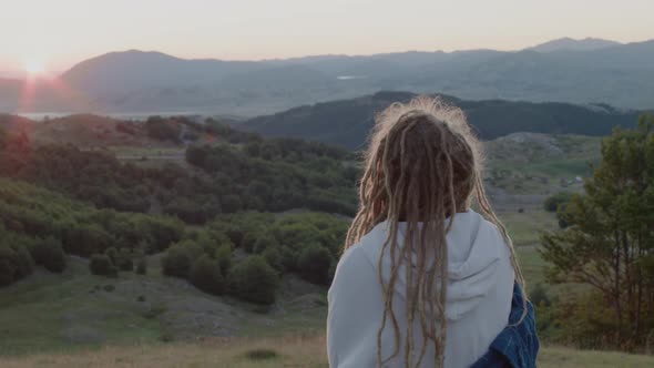 Back view of travel woman with dreadlocks near the tent standing on top of hill looking at sunrise. alt
