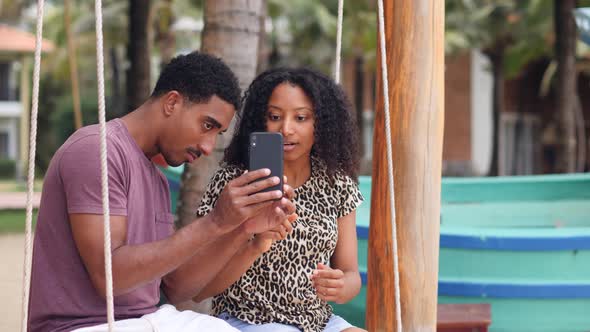 Lovely Young African Couple Sitting on a Swing and Using Smartphone alt
