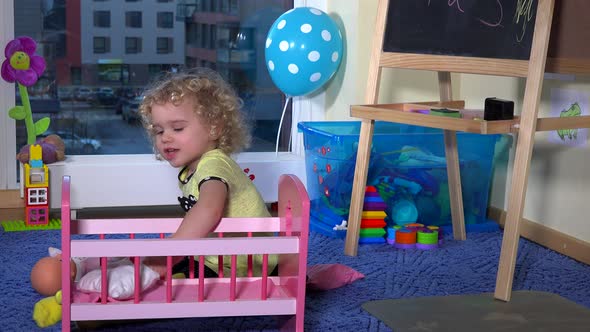 Sweet Blonde Toddler Girl Prepare Toy Bed Crib for Her Baby Doll at Home
