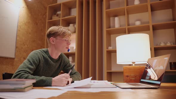 Young Man Studying at Home Having a Video Call on a Laptop Writing His Notes alt