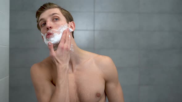 Positive Attractive Young Guy Applies Helium Foam in Front of the Mirror alt