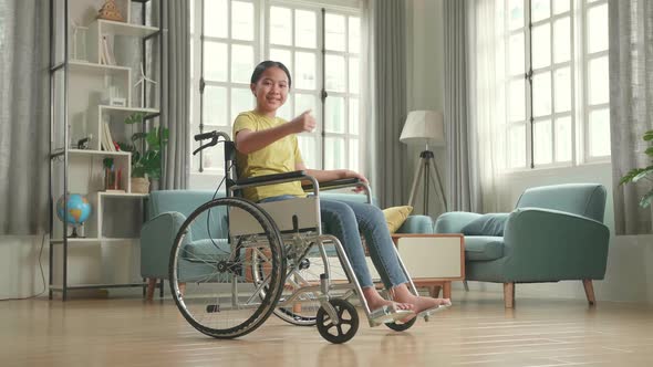 Asian Kid Girl Sitting In A Wheelchair Smiling And Showing Thumbs Up In Living Room alt