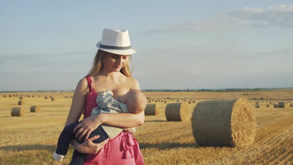 Stylish Young Mother in Hat Feeds the Baby Son with Breast in Hay Field alt