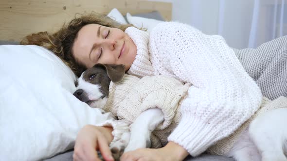 Smiling Woman Sleeping With Her Dog In Bed Wearing Cozy Knitted Sweaters.