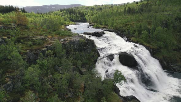 Aerial View Of Drone Operator Standing On Edge Of Cascading Waterfall Surrounded By Green Wilderness alt
