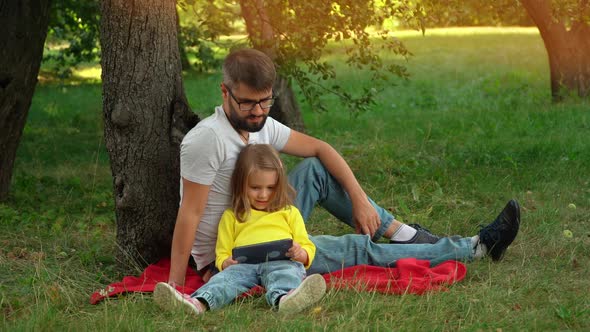 Father and Little Girl Sit on Lawn in Summer Park at Sunset with Tablet