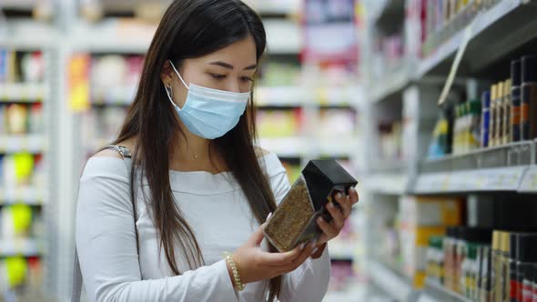 Asian woman in mask reading label of instant coffee at store alt